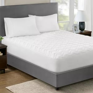 Home Design Classic Mattress Pads feature a quilted pattern, providing an extra layer of comfort and protection for mattresses. They have a sleek, white design and are shown on a bed with a grey upholstered headboard.