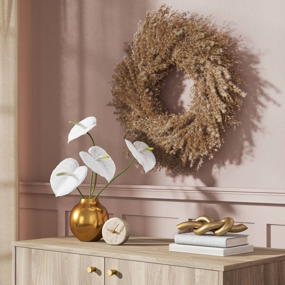 A wreath of dried grass hangs on a pink wall above a wooden cabinet with gold knobs. The cabinet holds a gold vase with white anthuriums, a circular beige clock, and a stack of books topped with a gold knot sculpture.