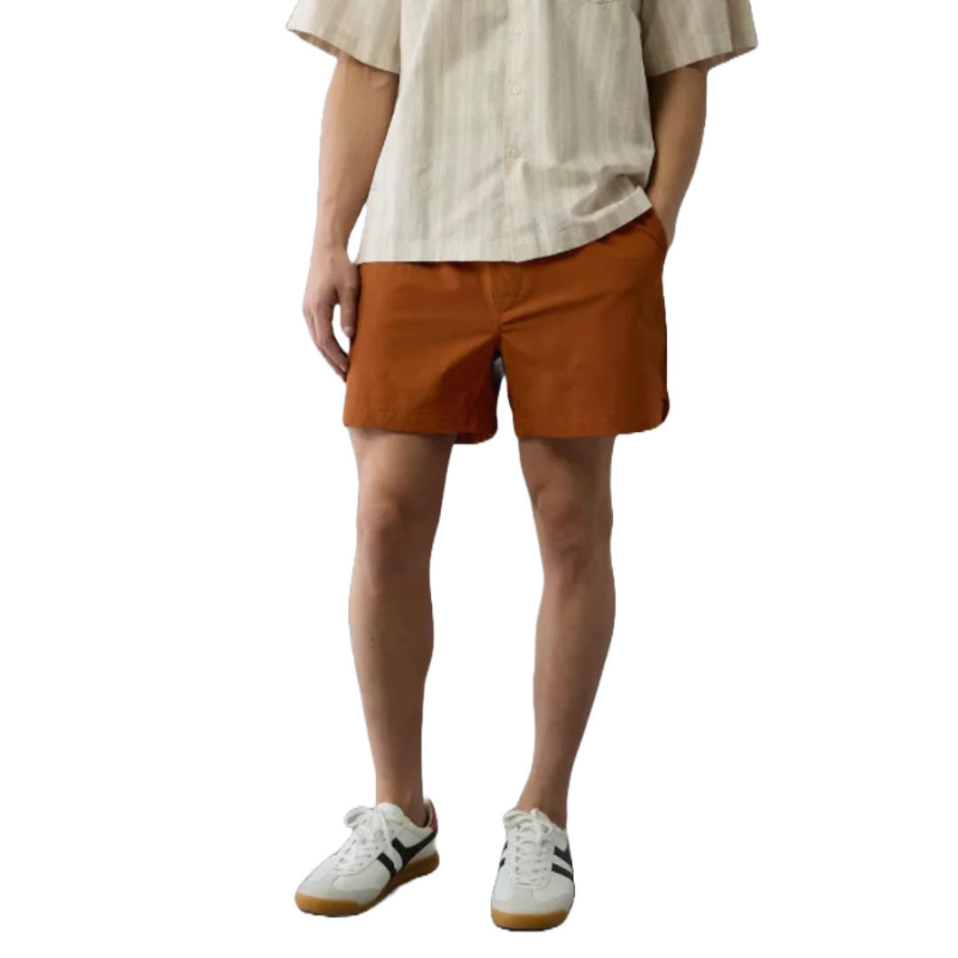 A person wearing rust-colored Flex Hybrid Shorts with a relaxed fit, paired with a light striped shirt and white sneakers featuring black detailing.
