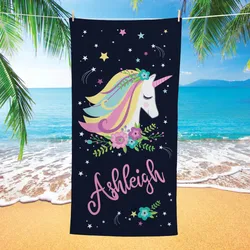 A beach towel featuring a colorful unicorn design surrounded by flowers and stars, with the name \“Ashleigh\“ written in cursive. Set against a beach backdrop with palm trees and ocean waves.
