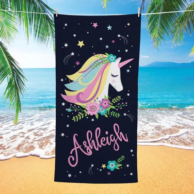 A beach towel featuring a colorful unicorn design surrounded by flowers and stars, with the name \“Ashleigh\“ written in cursive. Set against a beach backdrop with palm trees and ocean waves.