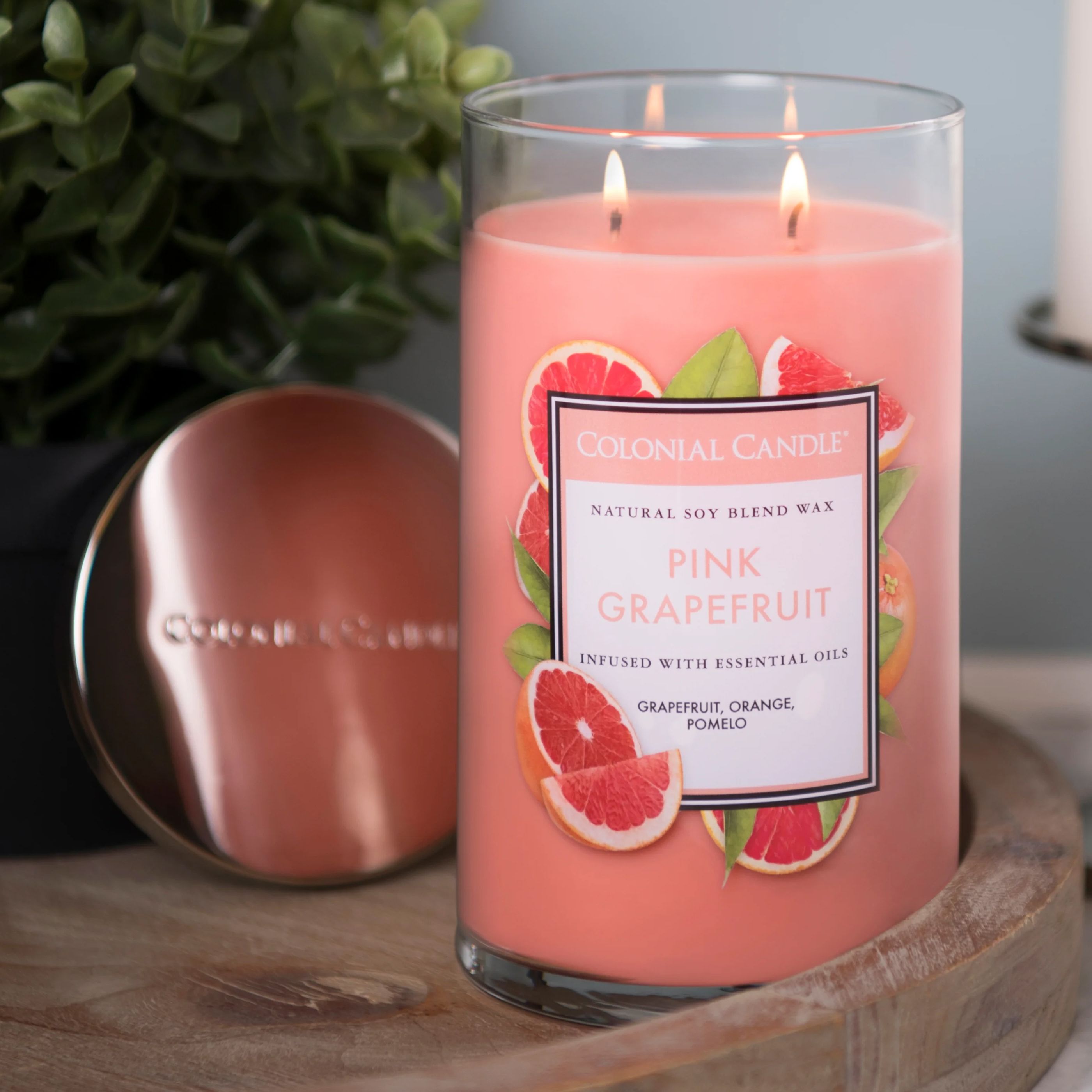 A pink grapefruit-scented Colonial Candle with natural soy blend wax, infused with essential oils like grapefruit, orange, and pomelo, displayed with its lid beside it on a wooden surface.