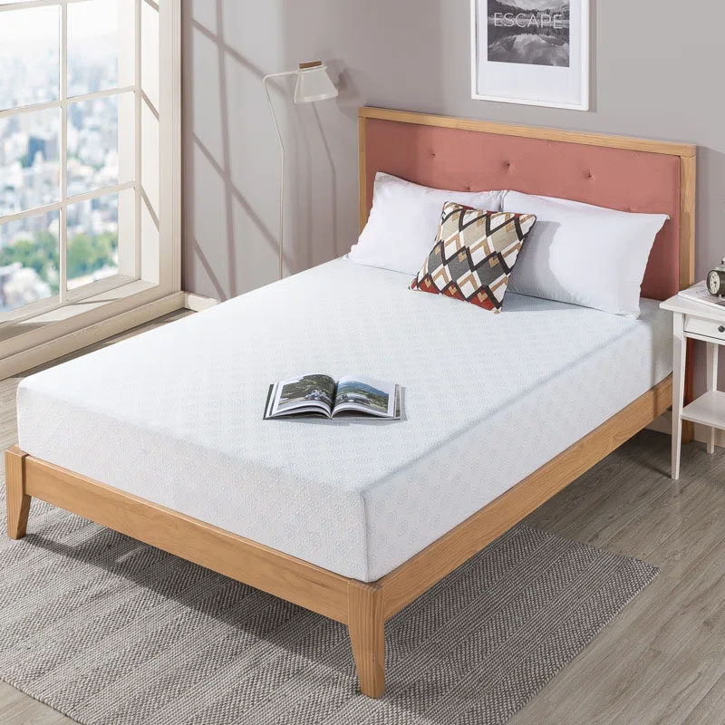 10\“ Queen Memory Foam Mattress on a wooden bed frame with a light pattern, accompanied by white pillows and a decorative cushion, beside a small white nightstand with a lamp in a bright room with large windows.