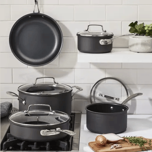 A set of non-stick cookware, including a frying pan, saucepans, and a stockpot, with glass lids and stainless steel handles, is displayed on a kitchen stove and shelves against a tiled backsplash.