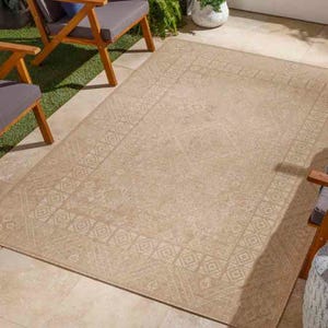 A beige 5' x 7' indoor/outdoor area rug with a subtle geometric border pattern, placed on a patio with beige tiles and surrounded by outdoor furniture, including wooden chairs with gray cushions and a potted plant in the background.