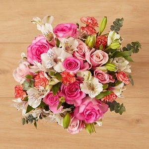 A bouquet featuring pink roses, white lilies, and red carnations with green foliage arranged on a wooden surface.