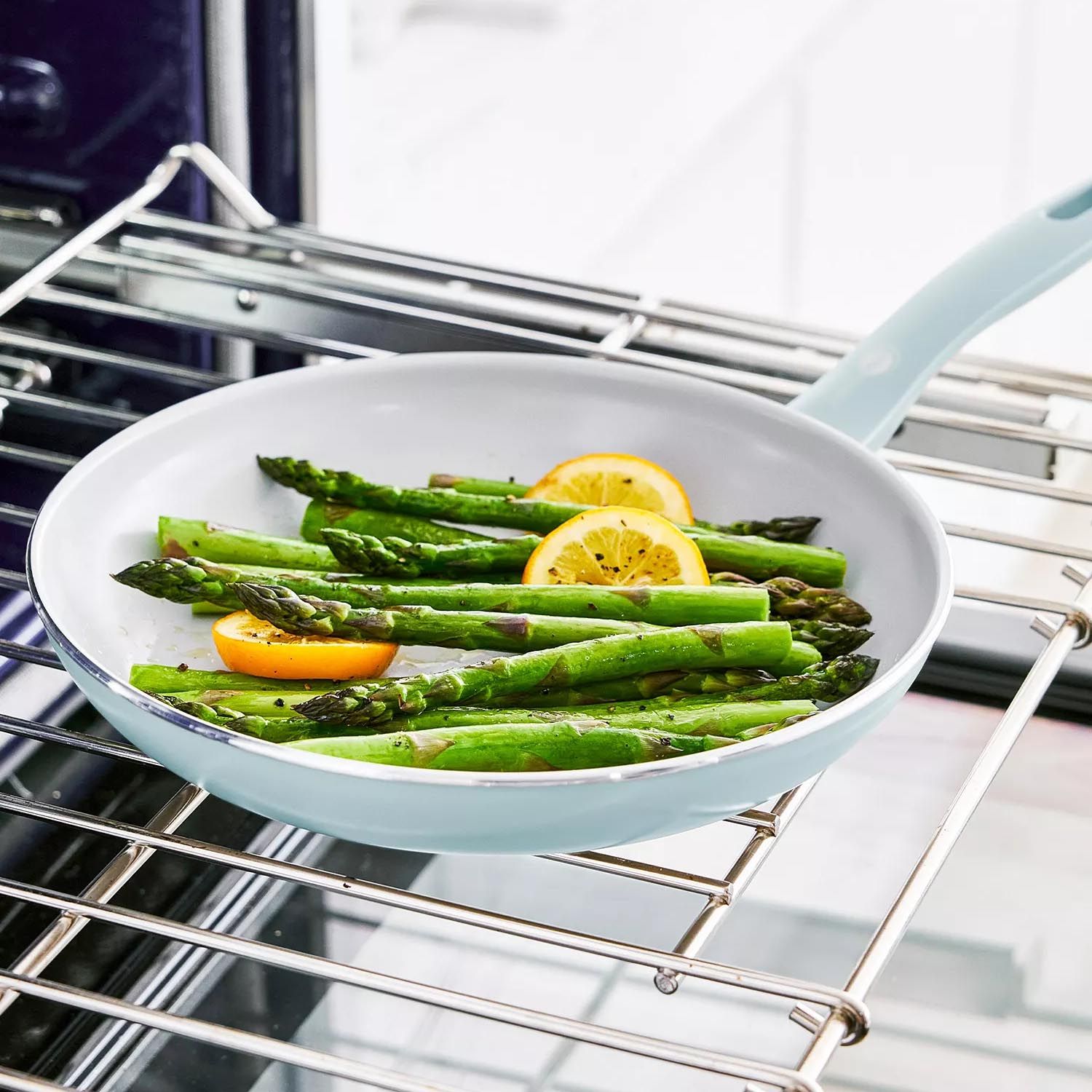 A light blue frying pan with asparagus and lemon slices is positioned on an oven rack.