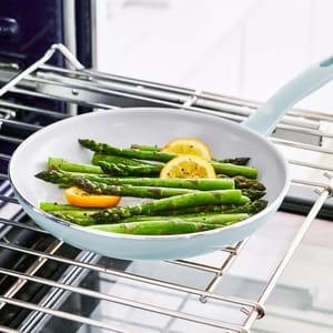 A light blue frying pan with asparagus and lemon slices is positioned on an oven rack.