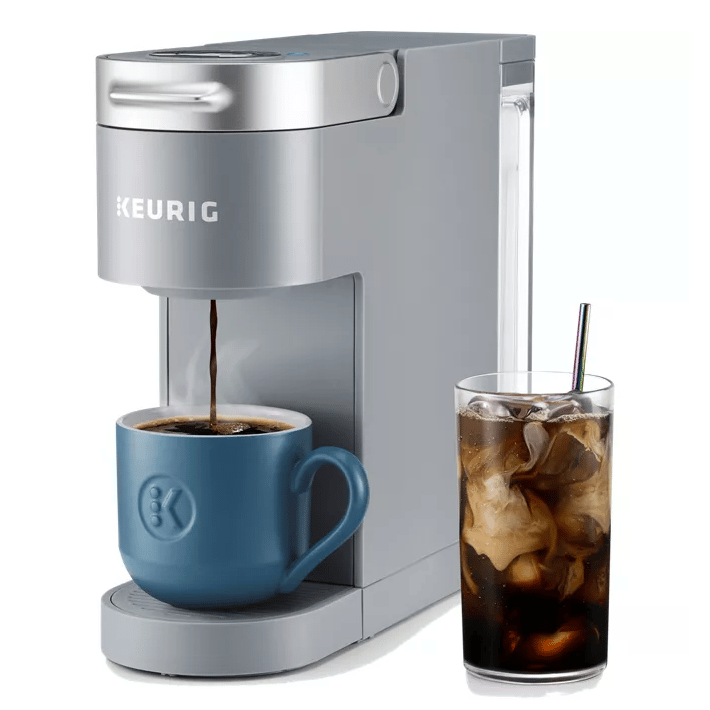 A Keurig coffee maker is dispensing hot coffee into a blue mug, with a tall glass of iced coffee and a straw beside it.