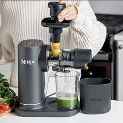 A person is using a Ninja juicer to extract juice from fruits and vegetables. The juicer is positioned on a countertop with a collection jug and pulp container.