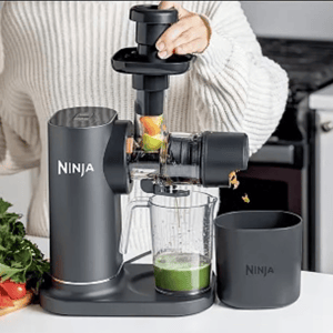 A person is using a Ninja juicer to extract juice from fruits and vegetables. The juicer is positioned on a countertop with a collection jug and pulp container.