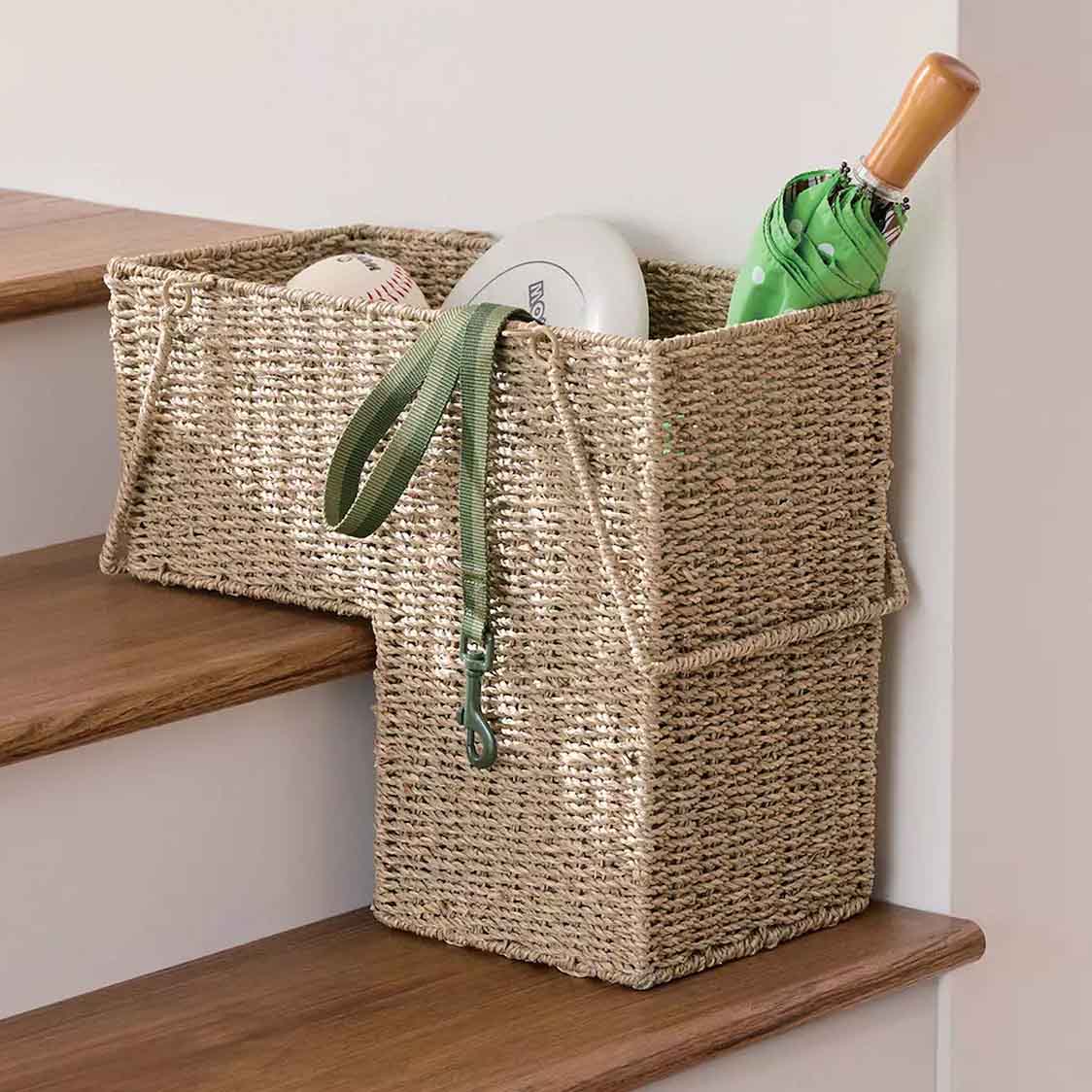A woven stair basket with handles sits on the stairs, designed to fit snugly over the step. It holds a baseball, frisbee, green umbrella, and a green dog leash, offering convenient storage for small items.