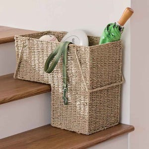 A woven stair basket with handles sits on the stairs, designed to fit snugly over the step. It holds a baseball, frisbee, green umbrella, and a green dog leash, offering convenient storage for small items.