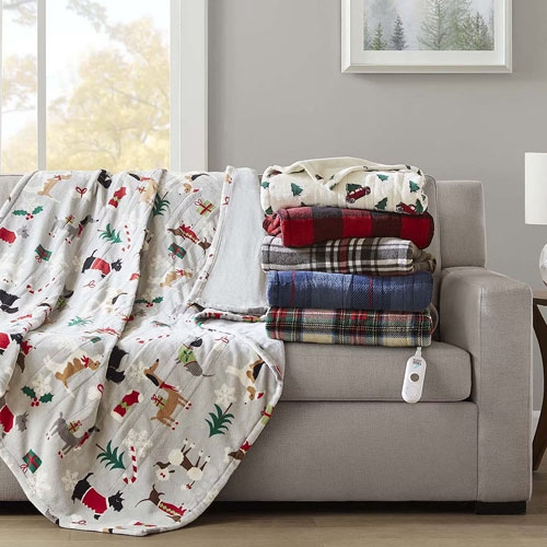 A pile of colorful blankets is stacked on a beige sofa, next to a white blanket with a festive holiday pattern and an electric blanket controller. The room features a window with fall foliage and a wall picture of trees.