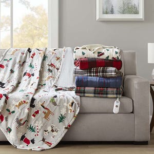 A pile of colorful blankets is stacked on a beige sofa, next to a white blanket with a festive holiday pattern and an electric blanket controller. The room features a window with fall foliage and a wall picture of trees.