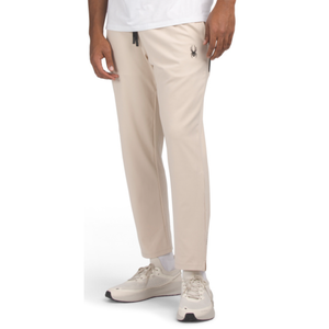Beige Spyder joggers featuring a small black spider logo on the upper thigh, styled with a black drawstring. Paired with white sneakers and a white top.