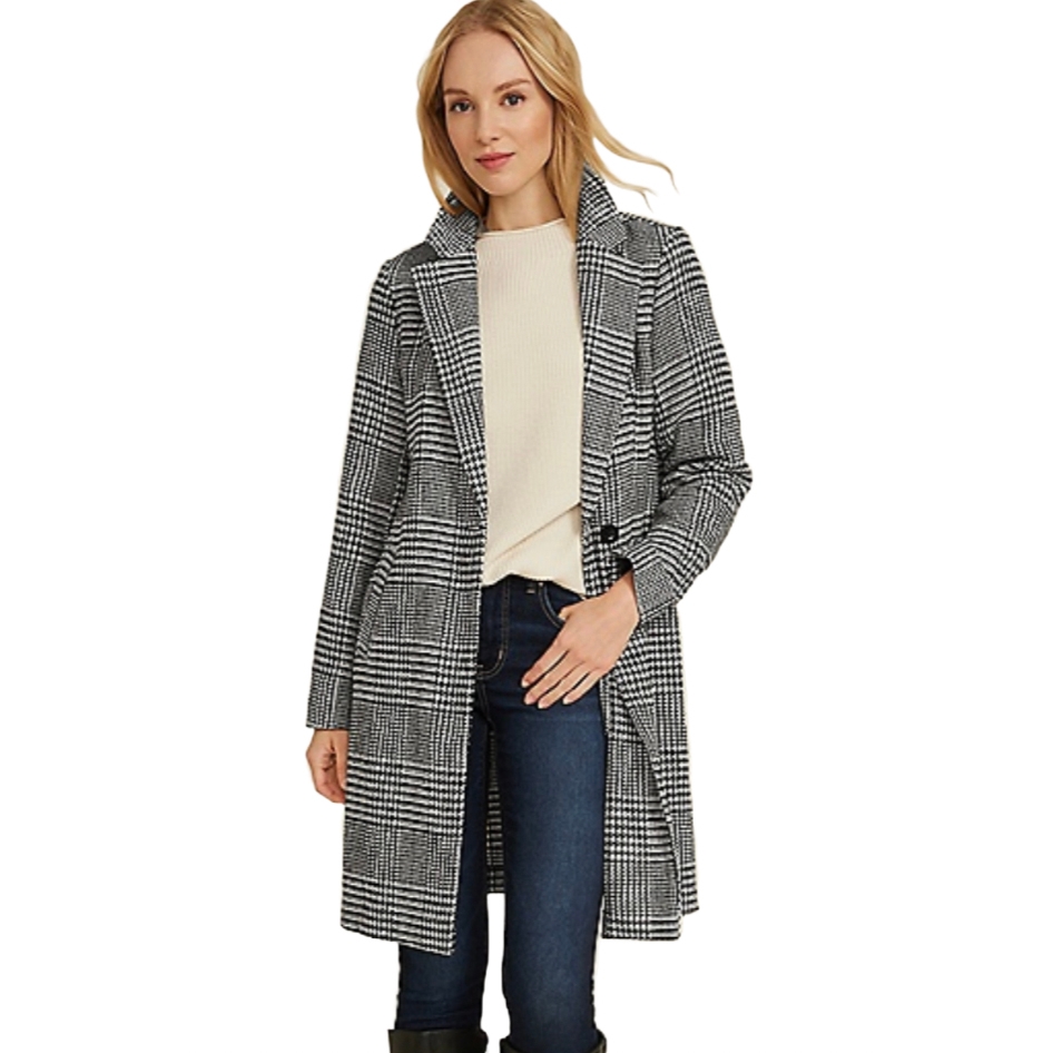 A woman wears a long houndstooth-patterned dress coat with a black and white color scheme. The coat features a notched lapel collar and is paired with dark jeans and a cream sweater.