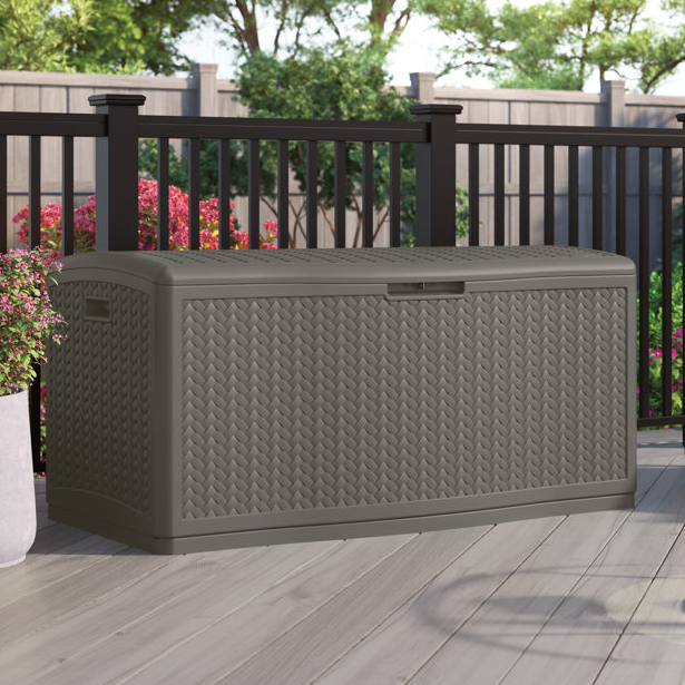 Outdoor storage box with a woven design, set on a patio with flowers and a fence in the background.