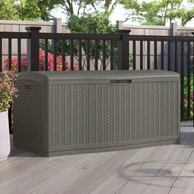 Outdoor storage box with a woven design, set on a patio with flowers and a fence in the background.