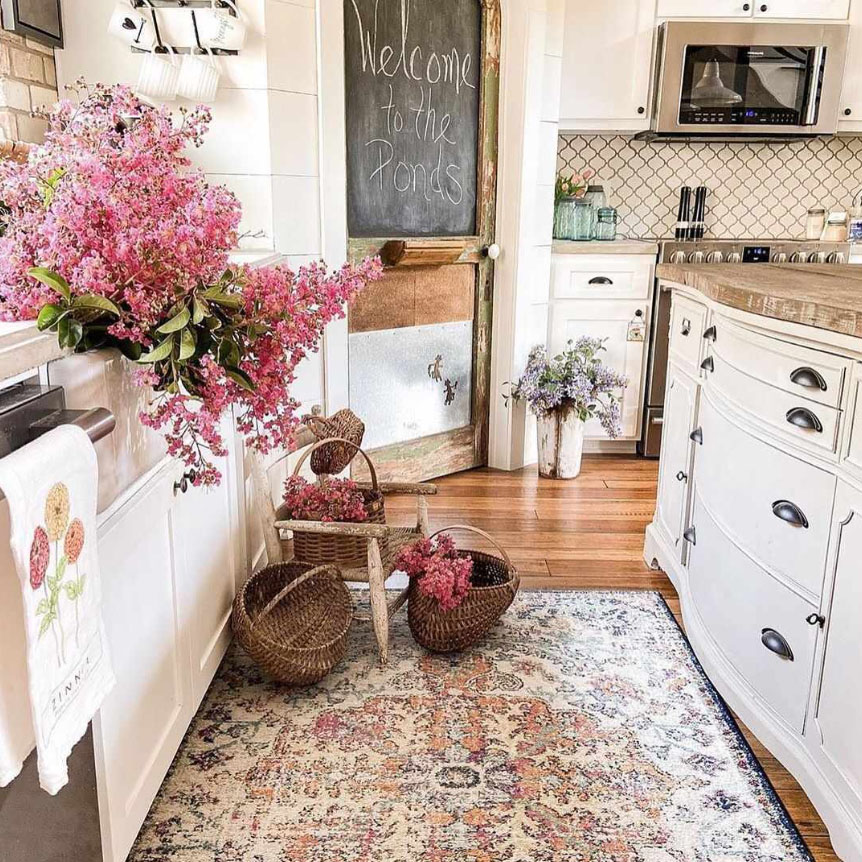 A cozy kitchen features white cabinetry, a wooden countertop, pink and purple floral arrangements, wicker baskets, and a decorative rug with pastel patterns. A chalkboard door says \“Welcome to the Ponds.\“