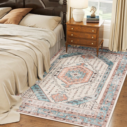 A bedroom scene featuring a bed with beige linens, a wooden nightstand with a lamp and a decorative item, and a patterned rug with blue, orange, and cream accents.