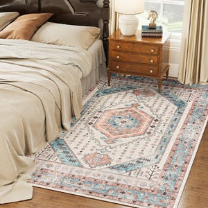 A bedroom scene featuring a bed with beige linens, a wooden nightstand with a lamp and a decorative item, and a patterned rug with blue, orange, and cream accents.