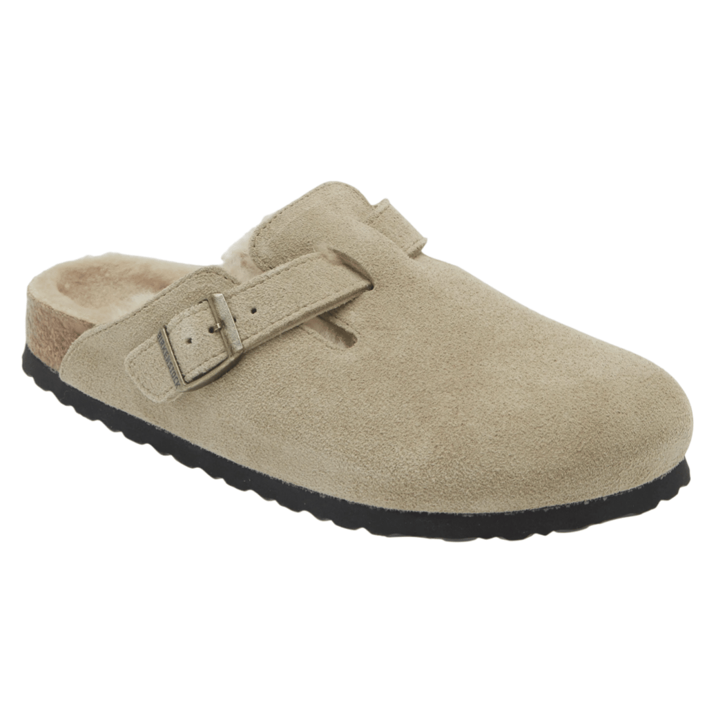 A tan suede clog-style slipper with a single adjustable strap and buckle; features a black rubber sole and shearling lining for comfort.