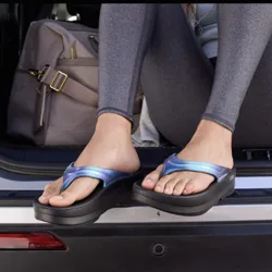 A person is sitting in the back of a vehicle wearing shiny blue flip-flops with black soles. They are dressed in gray leggings, and a beige bag is placed beside them.