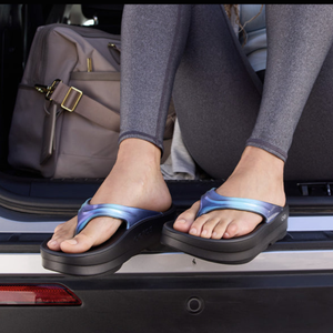 A person is sitting in the back of a vehicle wearing shiny blue flip-flops with black soles. They are dressed in gray leggings, and a beige bag is placed beside them.