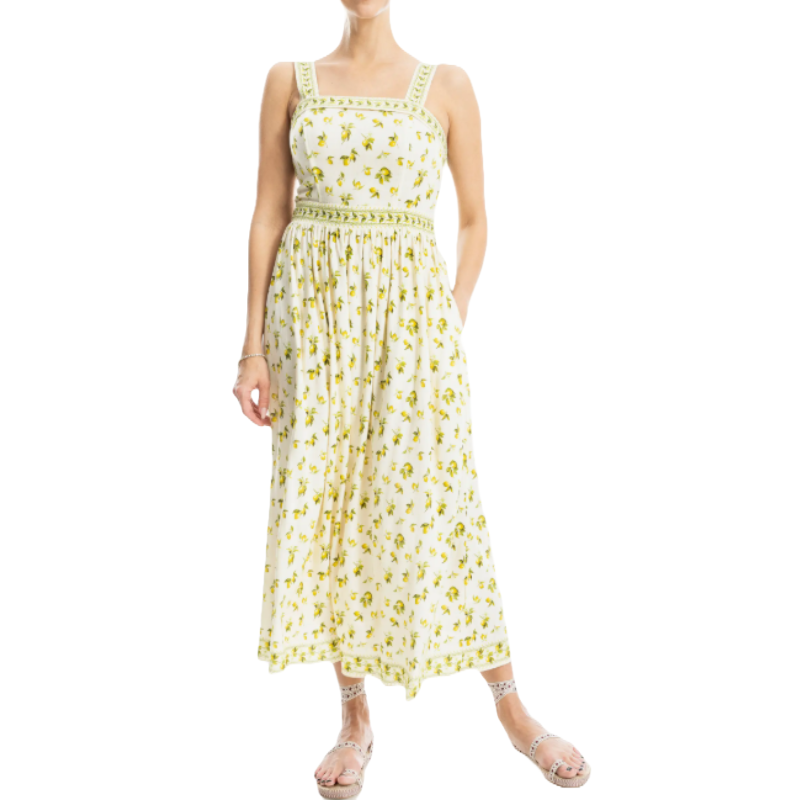 White flare maxi dress with yellow floral prints, featuring a square neckline, wide shoulder straps, and decorative green trim on the bodice and waist. Paired with studded sandals.
