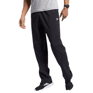 Reebok Training Essentials Pants are black athletic pants with a relaxed fit, featuring the Reebok logo on the upper thigh. They are paired with a grey shirt and black and white sneakers.