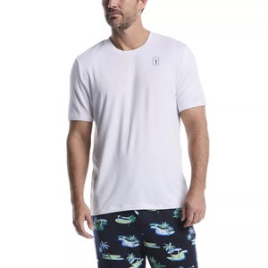 A man wears a plain white T-shirt with a small logo on the chest and dark shorts featuring palm tree and hammock patterns.
