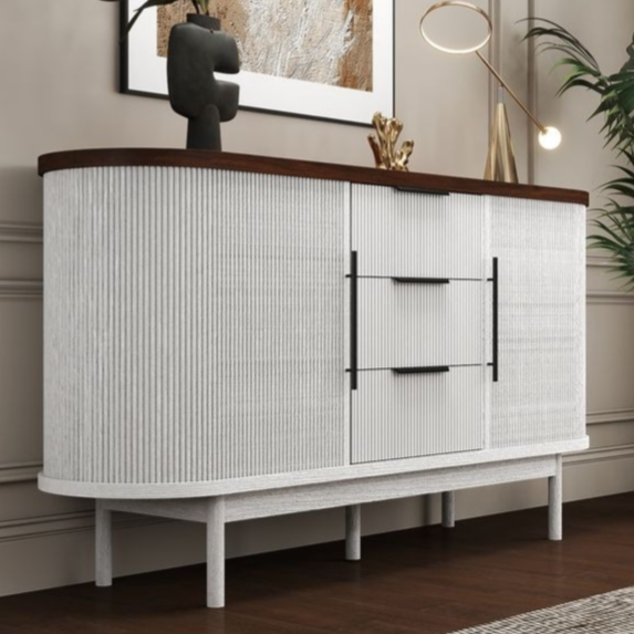 A modern sideboard features a white textured design with a dark wood top, decorated with a vase, abstract sculpture, and a small table lamp. It is set against a beige wall with a large framed artwork and accompanied by a tall potted plant.
