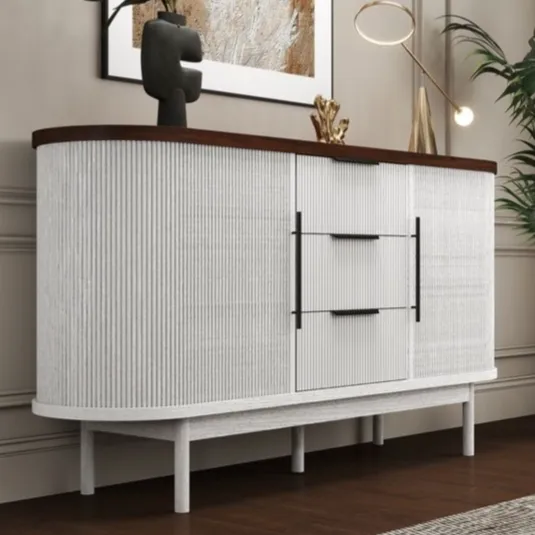 A modern sideboard features a white textured design with a dark wood top, decorated with a vase, abstract sculpture, and a small table lamp. It is set against a beige wall with a large framed artwork and accompanied by a tall potted plant.