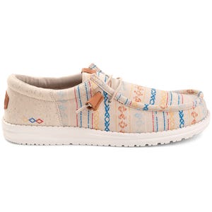A casual slip-on shoe with a colorful, woven pattern featuring stripes and geometric designs in blue, orange, and red hues on a light tan background. The shoe has a white sole and lace details.