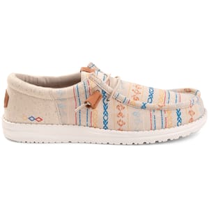 A casual slip-on shoe with a colorful, woven pattern featuring stripes and geometric designs in blue, orange, and red hues on a light tan background. The shoe has a white sole and lace details.