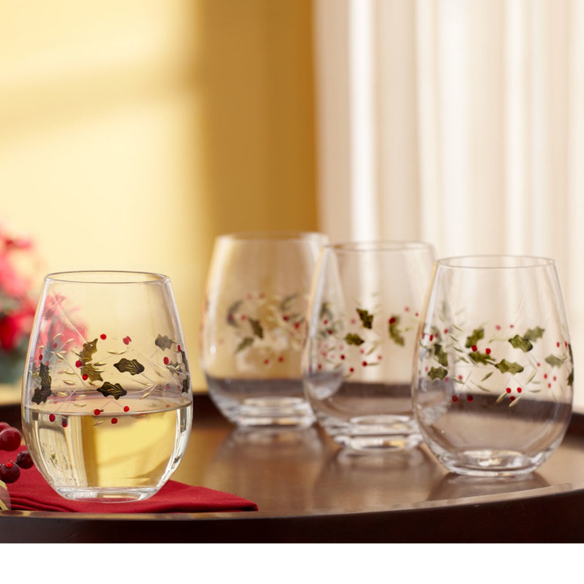 Four stemless wine glasses with holly and berry designs are displayed on a table, creating a festive appearance.