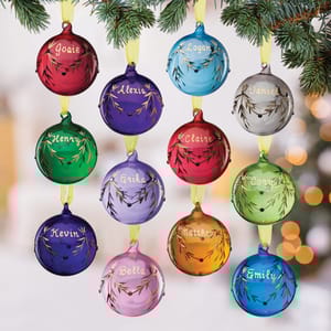 Colorful glass ornaments, each with a unique name and color representing a birthstone, are hanging on a pine branch. Names include Josie, Logan, Henry, Daniel, and others, with colors like red, blue, green, and more.
