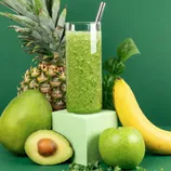 A green smoothie surrounded by a pineapple, mango, avocado, banana, spinach, and a green apple, with a metal straw in the glass.