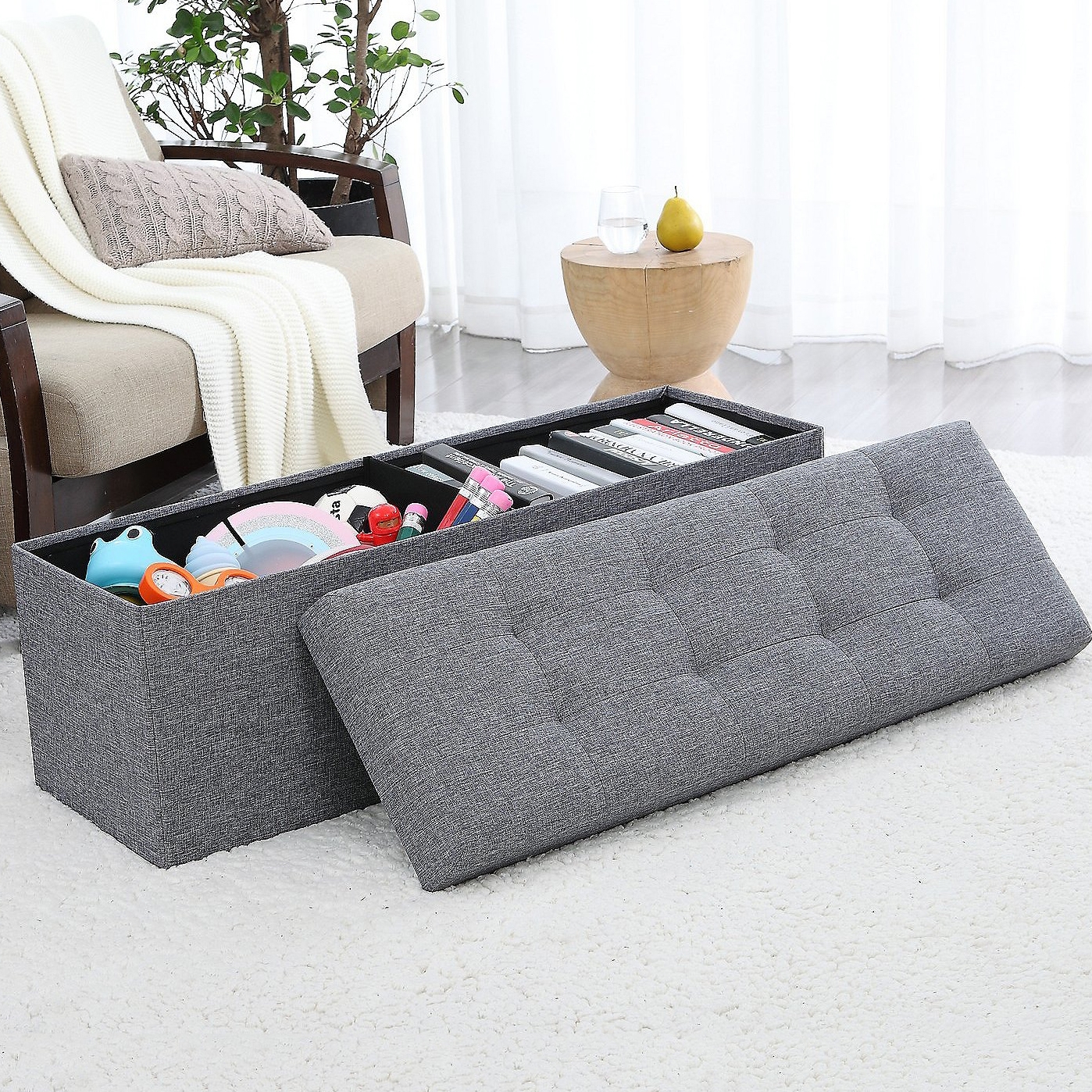 Gray tufted storage ottoman bench with a removable lid reveals organized compartments inside for storing various items like books and toys, placed on a white carpet near a chair and a small table.