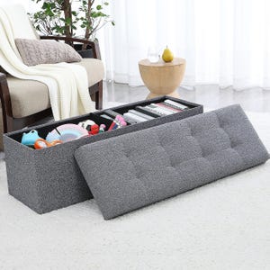 Gray tufted storage ottoman bench with a removable lid reveals organized compartments inside for storing various items like books and toys, placed on a white carpet near a chair and a small table.