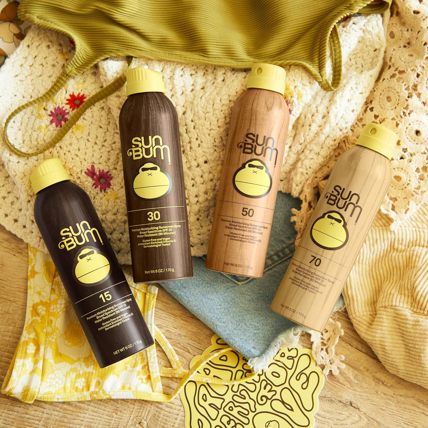 Sun Bum sunscreen spray bottles with SPF 15, 30, 50, and 70 are shown on a wooden surface, surrounded by beachwear and accessories.