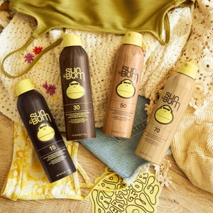 Sun Bum sunscreen spray bottles with SPF 15, 30, 50, and 70 are shown on a wooden surface, surrounded by beachwear and accessories.