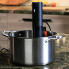 A stainless steel pot with handles, containing an immersion circulator or sous-vide cooker, is placed on a kitchen countertop.