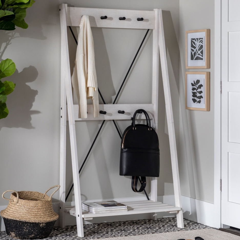 White freestanding coat rack with black hooks, beige sweater, black bag, and bottom shelf with books. Nearby are a woven basket, patterned floor, and framed wall art.