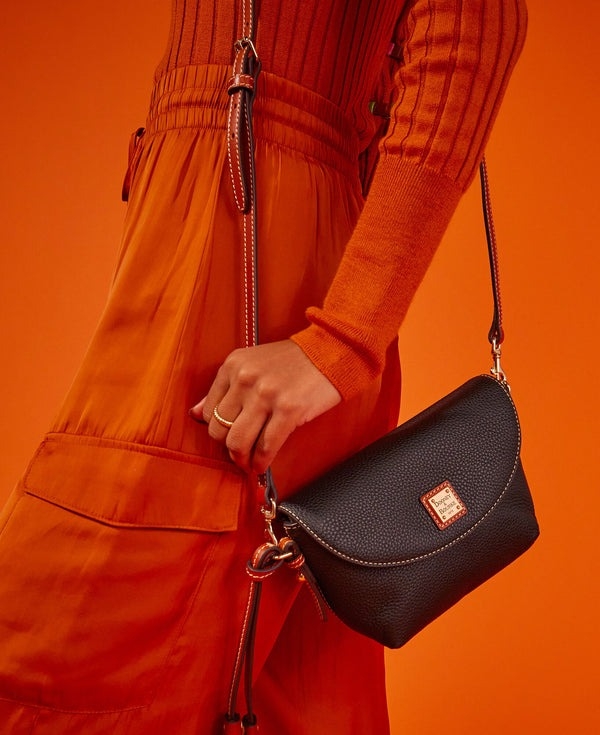 A person in an orange outfit holds a black crossbody bag with a textured surface and a small branded label.