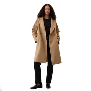 A person stands wearing a tan coat, black top, black trousers, and black loafers.