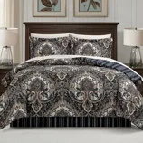 A bed set featuring a detailed paisley pattern with matching pillow shams, set against a dark wooden bed frame. Two table lamps with clear glass bases are on the nightstands beside the bed.
