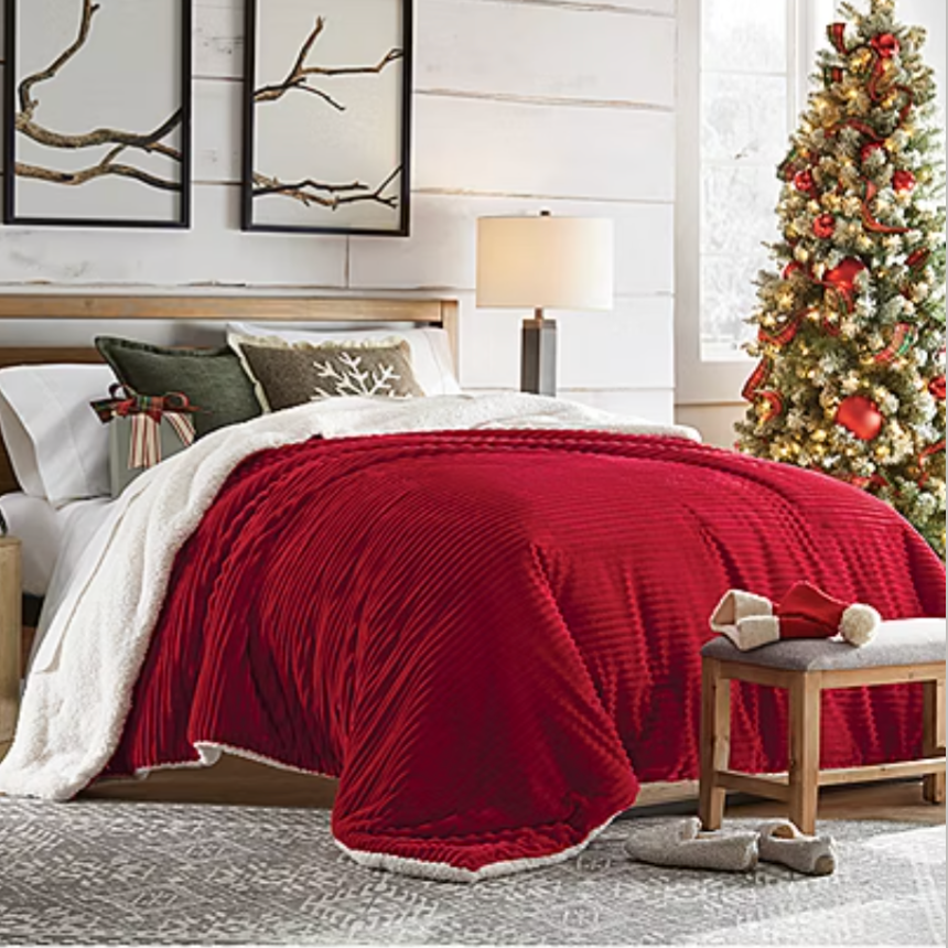 Bedroom with a wood bedframe, red textured blanket, white sheets, holiday-themed pillows, a side table with a lamp, a decorated Christmas tree, a wooden bench at the foot of the bed, and slippers on a patterned rug.