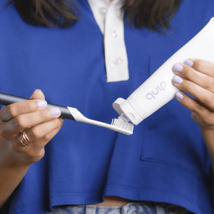 A person is applying toothpaste from a Quip tube onto a toothbrush, both held in their hands.